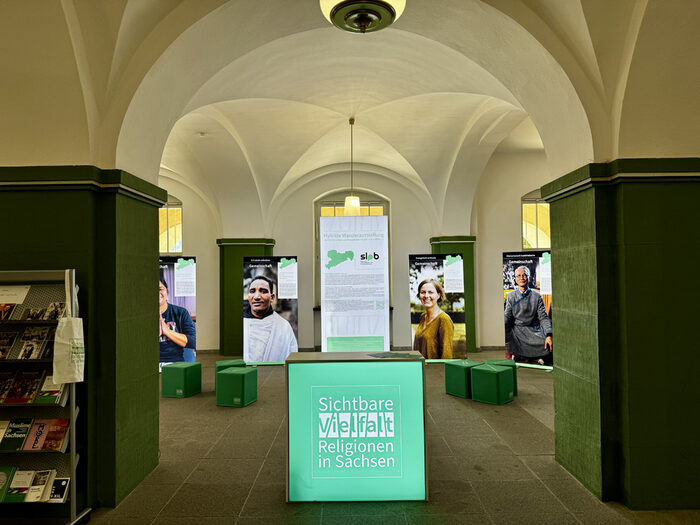 Blick in den Ausstellungsraum: Große Banner mit Fotos und Text stehen zwischen den historischen Bögen und Säulen.