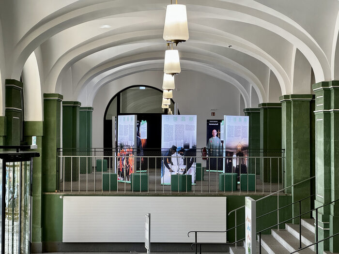 Blick in den Ausstellungsraum: Große Banner mit Fotos und Text stehen zwischen den historischen Bögen und Säulen.