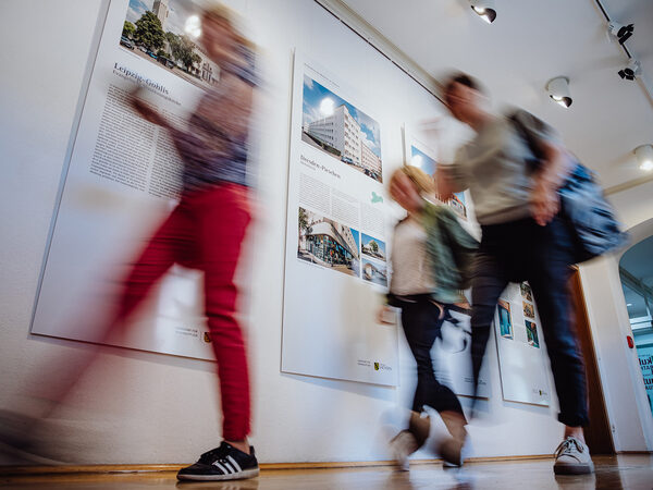 Tagungsteilnehmende eilen vorbei an einer Ausstellung zur Architektur der Moderne zum Tagungssaal