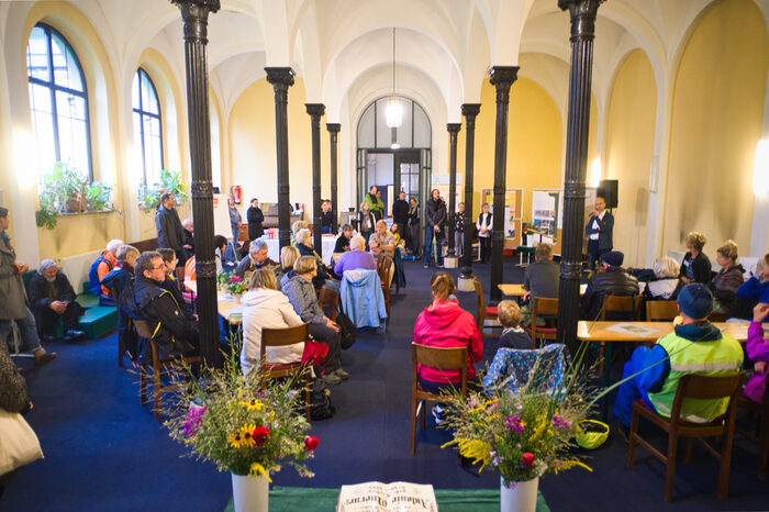 In der Feierhalle des Friedhofs sitzen Menschen auf Stühlen und hören einem Vortrag zu.