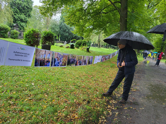 Eine Person mit Regenschirm schaut sic die Fotos der Veranstaltungen an. Die Fotos sind zwischen Bäumen an einer Leine befestigt.