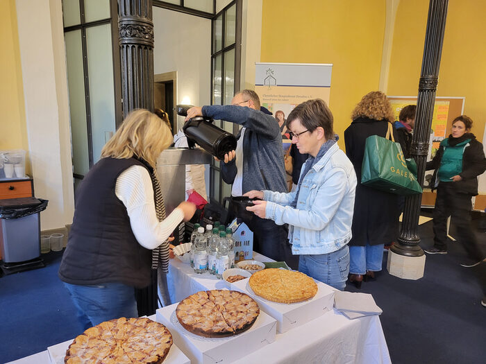 Tische mit Kuchen in der Feierhalle des Friedhofs; daneben stehen Menschen und unterhalten sich; im Hintergrund ein Poster