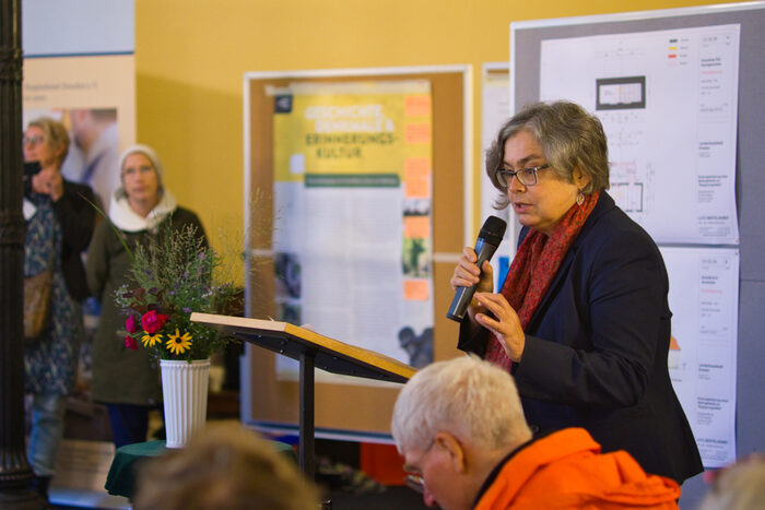 Eva Jähnigen spricht zu den Anwesenden. Sie benutzt ein Mikrofon. Im Hintergrund Plakatwände und einzelne Zuhörer.