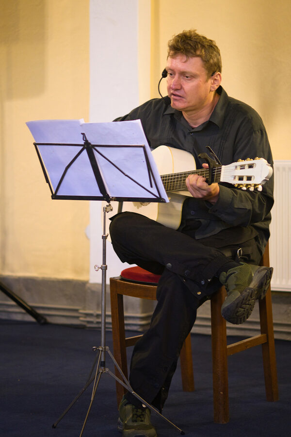 Markus Schröter sitzt auf einem Stuhl, spielt Gitarre und singt. Vor ihm ein Notenständer.