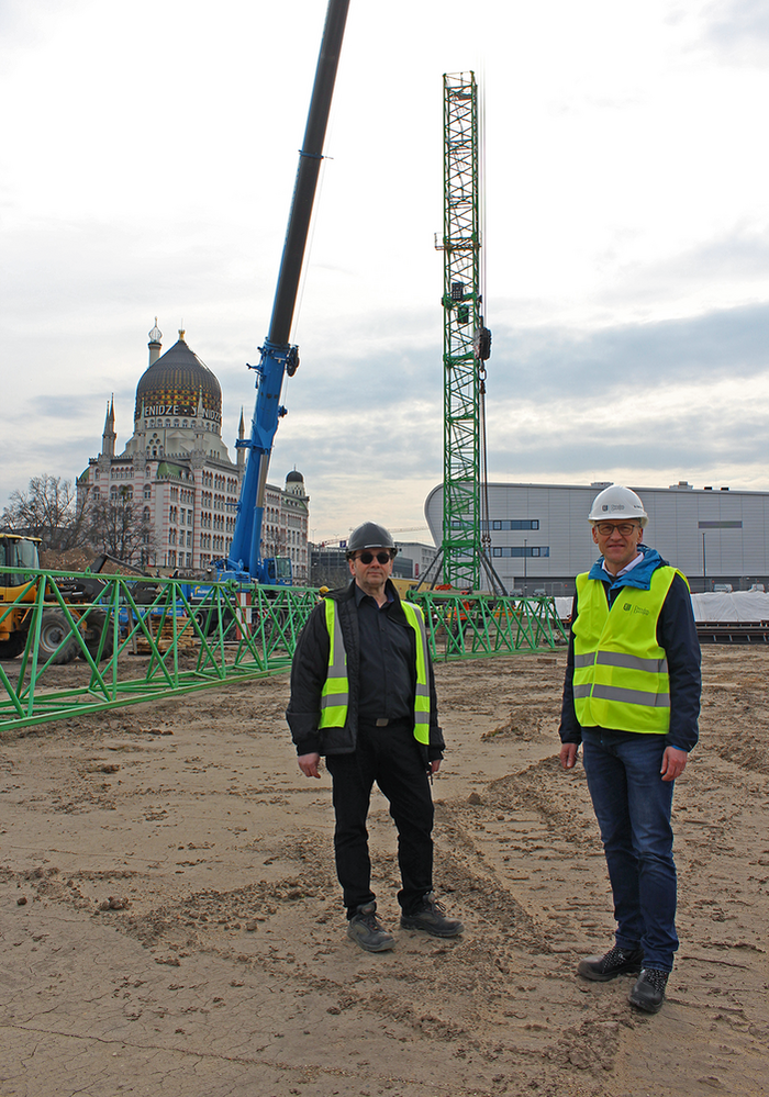 Zwei Männer mit Bauhelmen und gelben Warnwesten stehen auf einer Baustelle. Im Hintergrund wird ein großer Kran aufgebaut.