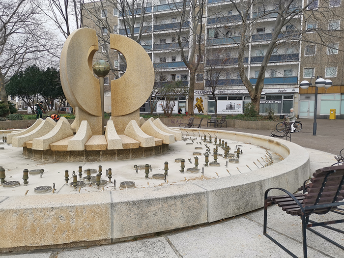 Foto von einem Brunnen ohne Wasser, dahinter eine Häuserzeile