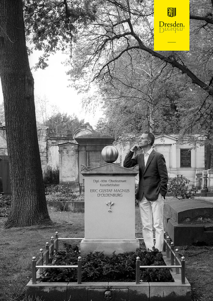 Ein Schwarz-Weiß-Foto, darauf ein Mann, der auf einem Friedhof an einem Grabmal lehnt. Oben auf dem Foto ist das gelbe Logo der Landeshauptstadt Dresden.