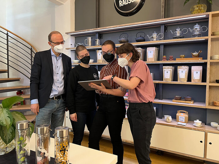 Vier Personen stehen in einem Laden und schauen auf ein Tablet. Im Regal im Hintergrund steht Teegeschirr.