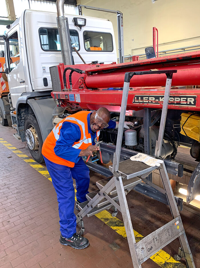 Ein Mann in blauer Arbeitskleidung arbeitet in einer Werkstatt an einem Absetzkipper. Vor ihm steht eine Leiter.