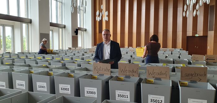 Mann steht vor Sammelbehältern und hält einen Stimmzettel