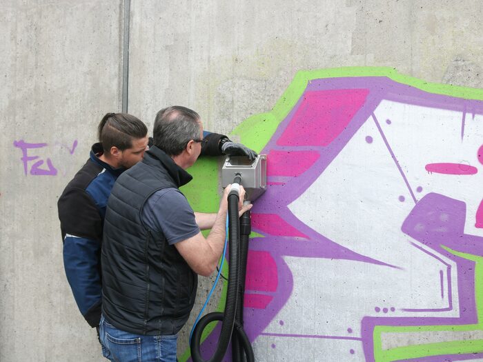 Zwei Männer stehen mit einem Arbeitsgerät an einer Wand und arbeiten