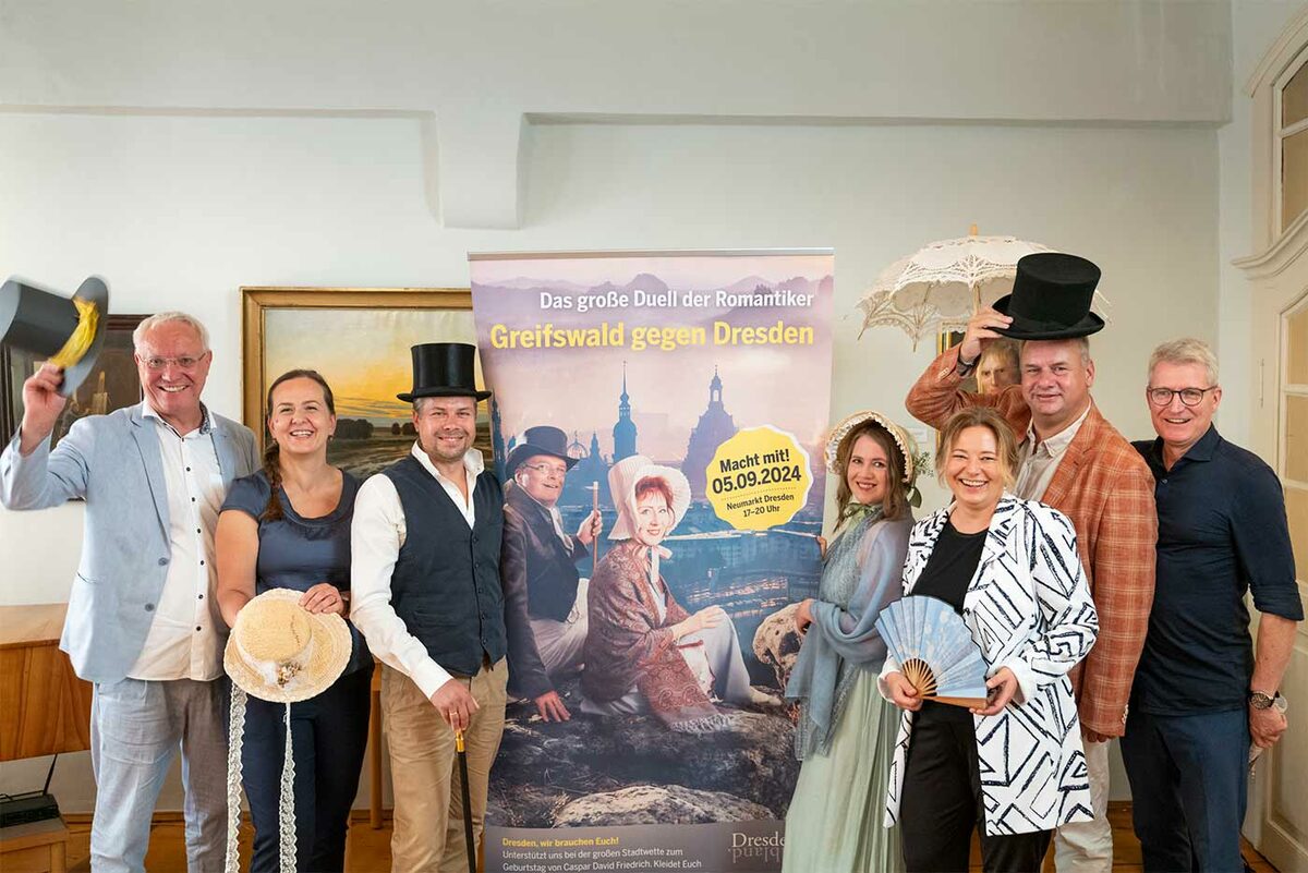 Männer und Frauen in historischen Gewändern mit Zylindern, Fächern, Sonnenschirmen neben einem Plakat, auf dem "Greifswald gegen Dresden"