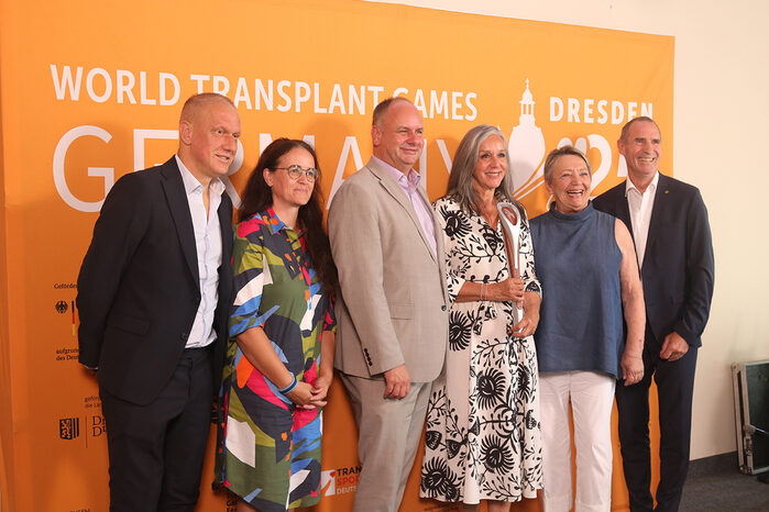 Drei Männer und drei Frauen vor einem orangefarbenen Plakat auf dem World Transplant Games steht