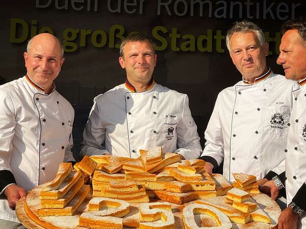 Vier Männer in weißer Jacke halten ein Kuchenblech mit Kuchen und vorn ist mit Kuchen die Zahl 250 gelegt