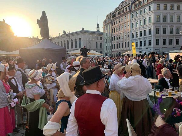 Viele Menschen mit Hüten und Zylindern und Kleidern und mit Bart und Weste stehen auf einem Platz um eine Statue herum. Die Sonne geht unter