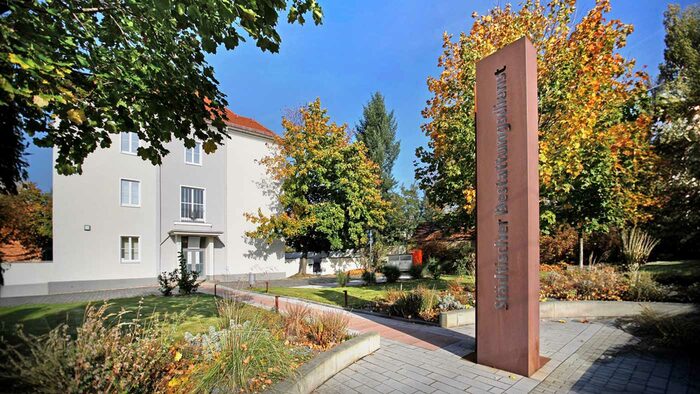 Hellgraues Gebäude umgeben von vereinzelten Bäumen und davor steht eine Stele mit der Aufschritt "Städtischer Bestattungsdienst"