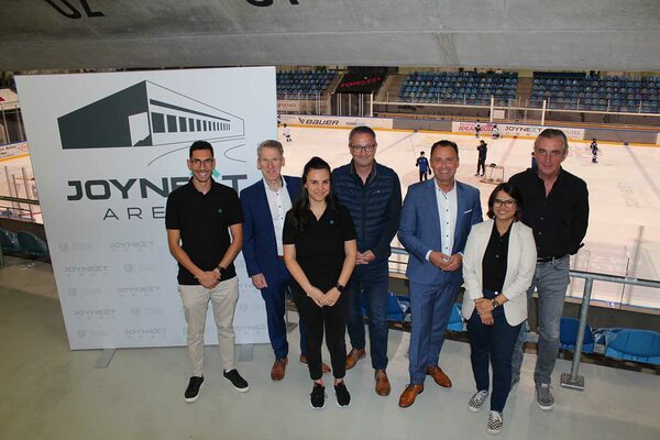 Fünf Männer und zwei Frauen stehen in einer Sporthalle und im Hintergrund spielen Menschen Icehockey