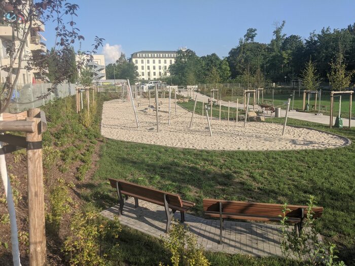 Das Bild zeigt einen Spielplatz mit einem Sandkasten und verschiedenen Klettergeräten. Im Hintergrund sieht man ein Gebäude und Bäume. Im Vordergrund befinden sich zwei Bänke.