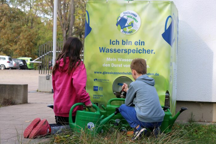 Zwei Kinder hocken vor einer Wasserbehälter mit zwei grünen Gießkannen