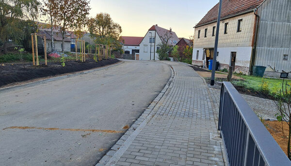 Straße mit Gehweg rechts und oben rechts steht ein Haus