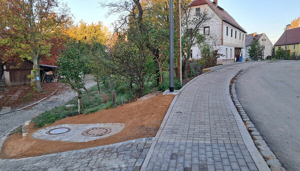 Gehweg in der Mitte des Bildes, links ein kleiner Weg und Bäume, weiter hinten vereinzelt Häuser, rechts Straße