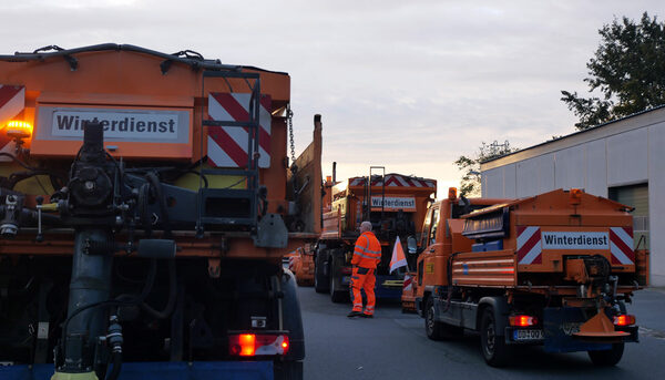 Drei orangefarbene Fahrzeuge mit der Beschriftung "Winterdienst" und in der Mitte ein Mann in organgefarbener Kleidung