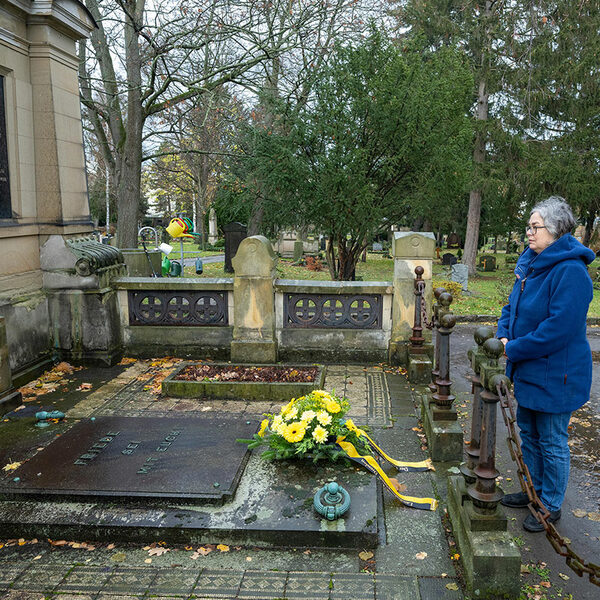 Grabstätte mit einer Mauer, in die ein Fenster eingelassen ist und eine Eule sitzt. Auf dem Grabstein liegt ein Kranz mit gelben Blumen liegt