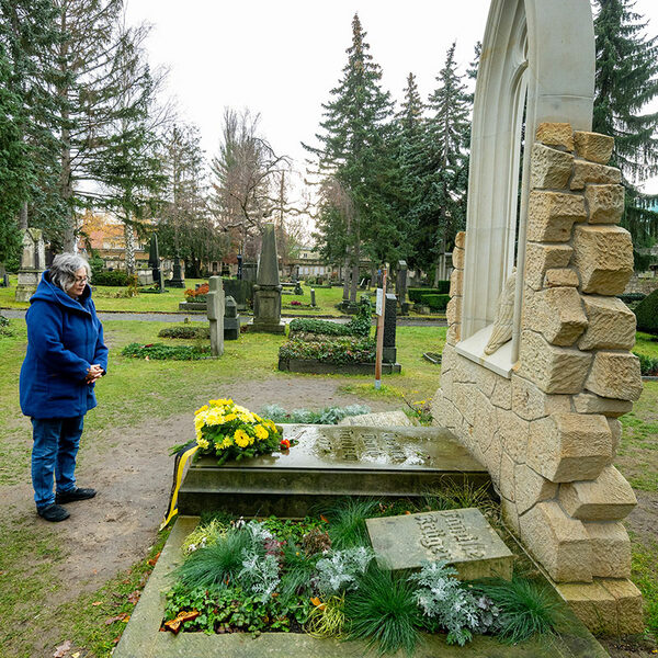 Frau mit grauen Haaren und Brille steht in blauer Jacke auf dem Friedhof vor einer Gruft, auf der ein Kranz mit gelben Blumen liegt