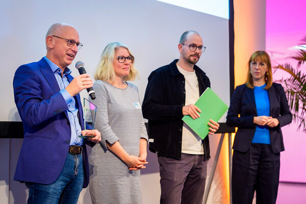 vier Personen stehen auf einem Podium, eine Person hält ein Mikrofon in der Hand und spricht.