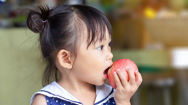 Kleines Mädchen mit schwarzen Haaren beißt gedankenverloren in einen roten Apfel