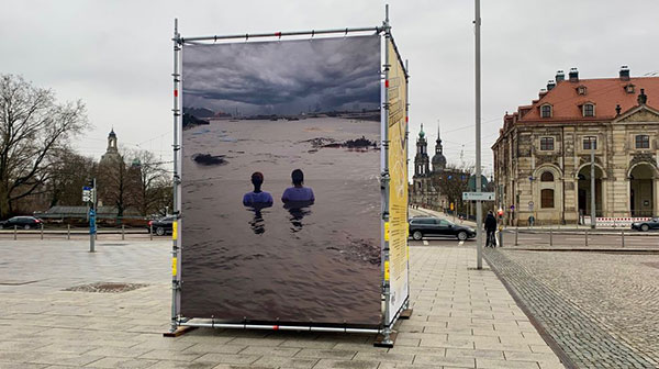 Vier Plakate rund herum auf einem Würfel auf einer Straße, das sichtbare Plakat zeigt zwei Menschen im Wasser