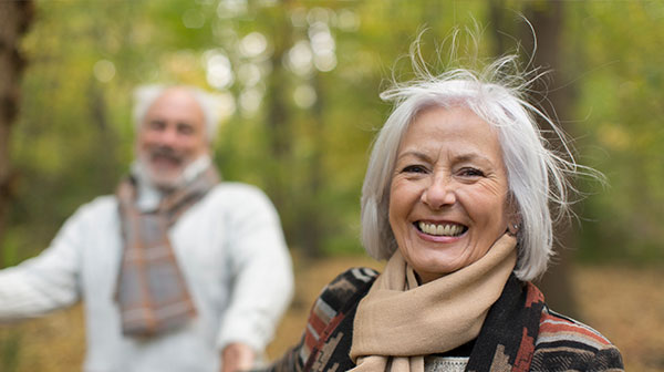 Frau mit grauen Haaren im Wald, braunes Tuch um den Hals, lächelnd, im Hintergrund verschwommen ein Mann mit grauen Haaren und buntem Schal