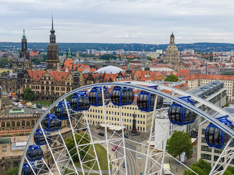 Luftaufnahme des Riesenrades „Wheel of Vision“ mit blau-weißen Gondeln auf dem Dresdner Postplatz. Im Hintergrund sind einige der Sehenswürdigkeiten wie der Zwinger und die Frauenkirche zu sehen.