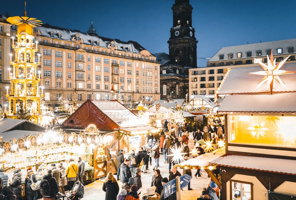 Blick auf den Striezelmarkt in Dresden. Alles ist festlich erleuchtet, Menschen schlendern an den geschmückten Buden vorbei. Links steht die weltweit größte erzgebirgische Stufenpyramide, mittig im Hintergrund sieht man die Kreuzkirche.